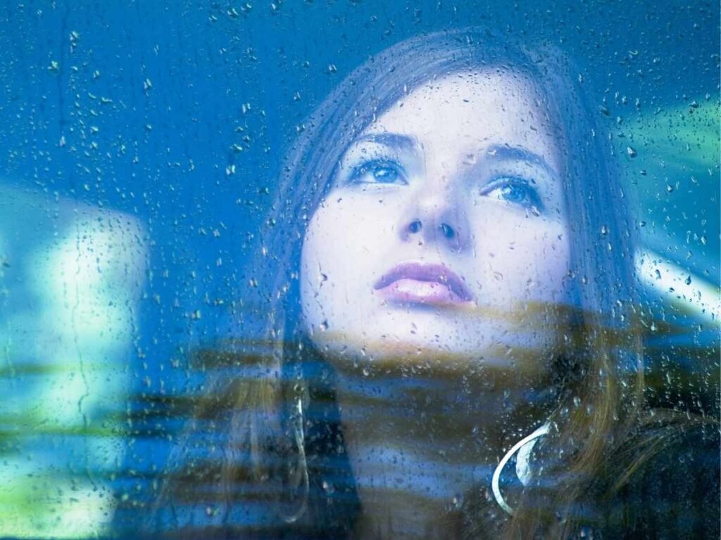 Woman looking thoughtfully through a rain-covered window, representing anxiety and emotional reflection